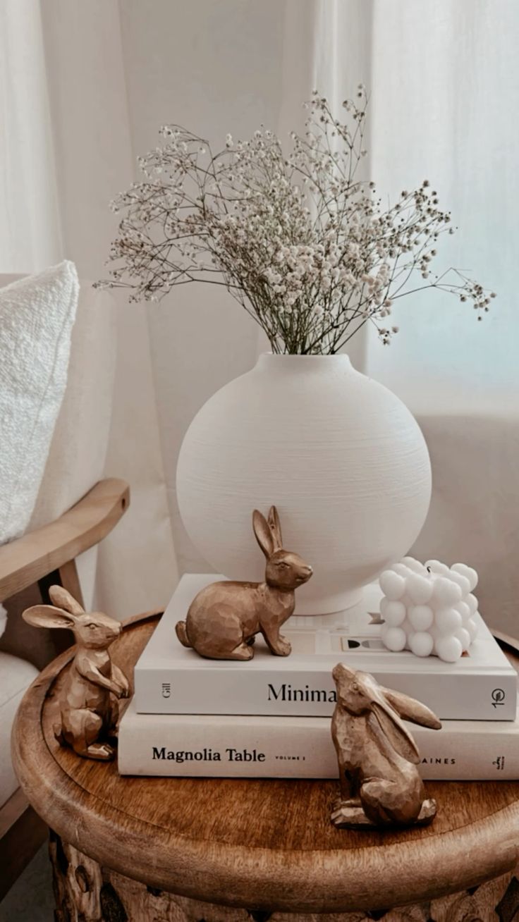 this picture contains a simple decor item ,a white vas with flowers in it, a white book and a gold easter bunny 